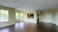 This single-floor space showcases an open-concept layout that seamlessly connects the main living area to the kitchen. Rich, dark hardwood flooring runs throughout, providing a striking contrast against the soft, neutral-toned walls and crisp white baseboards and ceiling.

The living area is generously sized, offering ample room for a variety of furniture arrangements. Two windows along the exterior wall invite natural light, brightening the interior and offering views of surrounding greenery. A track lighting fixture overhead provides additional illumination, ensuring the space remains well-lit throughout the day.

A defining architectural feature is the pass-through window that opens from the living area directly into the kitchen. The kitchen is equipped with dark cabinetry, stainless steel appliances including a refrigerator, and pendant lighting that adds a modern touch. This design element maintains visual connectivity between spaces while preserving a sense of definition.

Adjacent to the kitchen, a closet with built-in shelving offers practical storage. Overall, the space presents a clean, modern aesthetic with functional design choices that balance openness with organization.