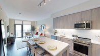 This single-floor space showcases a thoughtfully designed open-concept layout that integrates the kitchen, dining, and living areas into one cohesive environment. The kitchen is appointed with flat-panel, light wood-toned cabinetry that extends to the ceiling, offering generous storage. A white subway tile backsplash, stainless steel appliances including a built-in microwave and range, and an undermount sink contribute to the kitchen's clean, contemporary aesthetic. A white waterfall-edge island doubles as a dining surface, accommodating seating for multiple guests and serving as the natural focal point between the kitchen and living spaces. Track lighting overhead provides even illumination throughout. The living area is bathed in natural light streaming through a large window and glass door that opens to a private balcony, offering outdoor views and bringing a sense of airiness to the interior. Soft, neutral wall tones complement the warm wood finishes and light hardwood flooring, creating a welcoming and cohesive ambiance throughout the space. The overall design balances functionality with modern style.
