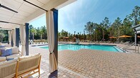 Located at 99 Athens Ct in Palm Coast, Florida, this single-story outdoor amenity facility serves as the centerpiece of the Pointe Grand community. The complex is anchored by a generously sized swimming pool with blue tile accents, complemented by an adjacent spa. The pool deck is finished with attractive paver stonework that extends throughout the entire outdoor area, providing a polished, resort-style aesthetic.

A covered pavilion runs along one side of the facility, featuring a ceiling fan, elegant white drape curtains, and comfortable outdoor seating arrangements including sofas and lounge chairs set atop area rugs. This shaded retreat offers a comfortable vantage point overlooking the pool. The pavilion structure incorporates large glass panel doors that connect the indoor and outdoor spaces seamlessly.

The surrounding sun deck is well-appointed with chaise lounges and umbrellas in a warm orange tone, providing ample space for sunbathing. An accessibility lift is present at the poolside, ensuring inclusive access for all residents. Recreational amenities such as a cornhole set bearing the Pointe Grand branding are available on the deck. The entire facility is framed by a backdrop of tall Florida pines and palm trees, enhancing the natural, tranquil ambiance of the space.
