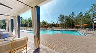 Located at 99 Athens Ct in Palm Coast, Florida, this single-story outdoor amenity facility serves as the centerpiece of the Pointe Grand community. The complex is anchored by a generously sized swimming pool with blue tile accents, complemented by an adjacent spa. The pool deck is finished with attractive paver stonework that extends throughout the entire outdoor area, providing a polished, resort-style aesthetic.

A covered pavilion runs along one side of the facility, featuring a ceiling fan, elegant white drape curtains, and comfortable outdoor seating arrangements including sofas and lounge chairs set atop area rugs. This shaded retreat offers a comfortable vantage point overlooking the pool. The pavilion structure incorporates large glass panel doors that connect the indoor and outdoor spaces seamlessly.

The surrounding sun deck is well-appointed with chaise lounges and umbrellas in a warm orange tone, providing ample space for sunbathing. An accessibility lift is present at the poolside, ensuring inclusive access for all residents. Recreational amenities such as a cornhole set bearing the Pointe Grand branding are available on the deck. The entire facility is framed by a backdrop of tall Florida pines and palm trees, enhancing the natural, tranquil ambiance of the space.