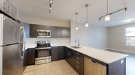 Located at 5418 W Black Hills Ln in Herriman, Utah, this well-appointed single-floor unit offers a clean, contemporary design throughout. The kitchen is a standout feature, showcasing dark espresso-toned cabinetry with sleek hardware, light quartz countertops, and a full suite of stainless steel appliances including a range, microwave, dishwasher, and refrigerator. A large peninsula with an integrated sink adds both functionality and style, while pendant lighting and track fixtures illuminate the space with a modern flair. Light wood-style flooring runs through the kitchen and several other rooms, contributing to a warm, cohesive aesthetic. The living areas are bright and neutral, with soft beige tones on the walls and plush carpet in select rooms. Multiple windows dressed with horizontal blinds invite natural light and offer views of the surrounding neighborhood. Bedroom spaces are generously sized, with ceiling fans and closet access adding practical comfort. The bathroom features tile flooring with a wood-grain appearance, white cabinetry with a rich wood-toned countertop, a backlit mirror, and a full soaking tub. The overall ambiance is clean, modern, and move-in ready, reflecting a thoughtfully designed living environment.