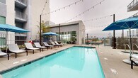 Located at 5401 Lankershim Blvd in North Hollywood, California, this outdoor amenity space offers a well-appointed, resort-inspired environment designed for relaxation and socializing. At its centerpiece is a generously sized swimming pool with crystal-clear blue water, flanked by rows of sleek white chaise lounges and shaded by blue market umbrellas. Overhead string lights add a warm, inviting ambiance that extends into the evening hours.

Adjacent to the pool, a spacious open-air lounge area features a built-in fire pit table surrounded by a large sectional sofa with vibrant, colorful accent pillows, creating a comfortable gathering spot. The outdoor kitchen station is equipped with a stainless steel built-in grill, stone countertops, and bar seating, making it ideal for outdoor entertaining.

The courtyard areas are thoughtfully landscaped with tropical palms, succulents, and lush greenery set within large concrete planters, contributing to a California-inspired aesthetic. Glass railings and clean-lined architectural finishes throughout the space reinforce a modern design sensibility. The multi-story residential building surrounding the deck features private balconies that overlook the amenity level, seamlessly integrating indoor and outdoor living.