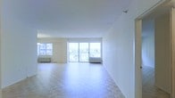 This single-floor space offers a generous, open-plan layout filled with abundant natural light. The main living area is defined by classic parquet flooring arranged in a herringbone-style pattern, adding warmth and character to the otherwise clean, minimalist interior. Bright white walls and a smooth white ceiling enhance the sense of airiness throughout.

A standout feature is the expansive wall of floor-to-ceiling sliding glass doors that leads directly to a private balcony. This opening floods the interior with daylight and provides unobstructed views of the surrounding greenery and neighboring buildings, creating a seamless connection between indoor and outdoor living. A secondary window to the left of the main doors further amplifies natural light in the space.

The layout is open and flexible, offering ample room for a variety of furniture arrangements. Baseboard heating units are visible beneath the windows, providing practical climate control. The overall ambiance is clean, airy, and bright, with a neutral palette that serves as a versatile backdrop for personalization.