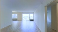 This single-floor space offers a generous, open-plan layout filled with abundant natural light. The main living area is defined by classic parquet flooring arranged in a herringbone-style pattern, adding warmth and character to the otherwise clean, minimalist interior. Bright white walls and a smooth white ceiling enhance the sense of airiness throughout.

A standout feature is the expansive wall of floor-to-ceiling sliding glass doors that leads directly to a private balcony. This opening floods the interior with daylight and provides unobstructed views of the surrounding greenery and neighboring buildings, creating a seamless connection between indoor and outdoor living. A secondary window to the left of the main doors further amplifies natural light in the space.

The layout is open and flexible, offering ample room for a variety of furniture arrangements. Baseboard heating units are visible beneath the windows, providing practical climate control. The overall ambiance is clean, airy, and bright, with a neutral palette that serves as a versatile backdrop for personalization.