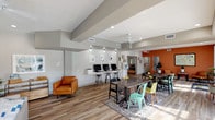 This single-floor community clubhouse and leasing office is thoughtfully designed to serve as a welcoming hub for residents and prospective tenants alike. The interior showcases wood-look plank flooring throughout, vaulted ceiling details with exposed beam accents, and a bright, neutral color palette punctuated by bold terracotta orange accent walls that lend warmth and energy to the space.

The main reception and leasing area features an open-plan layout with a dedicated workstation desk, seating for visitors, and direct access to private offices. A well-appointed administrative office, highlighted by its rich orange walls, houses workstations, a multifunction printer, and ample storage, providing a fully functional workspace for staff.

A comfortable lounge area includes a contemporary gray sofa, leather accent chairs, a ceiling fan, and curated artwork, creating an inviting retreat for residents. An adjacent co-working and social zone offers communal tables with colorful seating and a built-in computer workstation bar, ideal for productivity or casual gatherings.

A secondary waiting lounge provides navy blue seating and natural light through a large picture window. Sliding glass doors open directly to an outdoor pool area, seamlessly blending indoor and outdoor living. The overall ambiance is modern, vibrant, and community-focused.