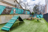 Located in Atlanta's upscale 30305 zip code, this ground-level outdoor amenity space is purpose-built for pets and their owners. The centerpiece is a generously sized, fully enclosed dog park surfaced with lush artificial turf, providing a clean and low-maintenance area for dogs to exercise and socialize year-round. The space is secured by sturdy black metal fencing with a gated entry, ensuring safety for all four-legged visitors.

The park is thoughtfully equipped with agility features, including ramp structures, natural log obstacles, and tunnel elements, offering mental and physical stimulation for dogs of all sizes. Waste disposal stations are conveniently positioned throughout the area. Playful branded signage lines the perimeter wall, adding a lively and welcoming atmosphere to the space.

Adjacent to the turf area is a covered composite wood deck, sheltered by the building's overhang and accented with warm wood-slat privacy screens. A classic bench seat provides a comfortable spot for owners to relax while supervising their pets. The multi-story building backdrop, open sky above, and surrounding greenery contribute to an inviting outdoor environment that balances functionality with thoughtful design.