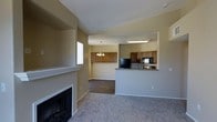 This single-floor residence is designed around an open-concept floor plan that seamlessly connects the living area and kitchen. The living room is carpeted and anchored by a white-mantled fireplace, adding a classic focal point to the space. Neutral beige walls throughout create a warm, cohesive atmosphere.

The kitchen is positioned in a raised, partially enclosed nook visible from the living area, featuring medium-toned wooden cabinetry, dark granite or laminate countertops, and black appliances including a refrigerator and built-in microwave. An overhead fluorescent fixture provides functional lighting above the workspace, while a decorative chandelier with warm-toned bulbs hangs nearby, adding a touch of elegance.

A pass-through opening connects the kitchen visually to a secondary space — likely a dining area — defined by its hardwood-style flooring that contrasts with the carpeted living room. A window allows natural light to filter in, brightening the interior. The layout is practical and efficiently organized, making it well-suited for everyday living.