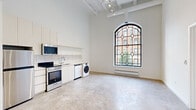 Located at 1048 W Lawrence Ave in Chicago's Uptown neighborhood, this single-floor unit blends historic architectural character with modern finishes. The space is immediately defined by its soaring ceilings with exposed white structural beams and industrial-style track lighting, lending a loft-like openness throughout.

A dramatic arched leaded-glass window serves as a stunning focal point, flooding the interior with natural light and offering views of the surrounding streetscape and mature trees. The polished concrete flooring extends seamlessly through the open living and kitchen area, enhancing the industrial aesthetic.

The kitchen is fitted with white cabinetry, a white tile backsplash, and a full suite of stainless steel appliances, including a range, refrigerator, built-in microwave, and dishwasher. An in-unit washer and dryer add everyday convenience. Baseboard heating runs beneath the windows throughout the unit.

The bedroom features its own arched leaded-glass window, continuing the architectural character of the main living area, and includes access to a dedicated walk-in closet with built-in shelving and rail storage. The bathroom is clean and functional, outfitted with a walk-in shower with a sliding glass door and a handheld showerhead, set on marble-tiled flooring.