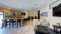This single-floor community space is thoughtfully designed to accommodate a variety of needs in a warm, inviting environment. The main lounge area features vaulted ceilings with ceiling fans, large tile flooring, and abundant natural light streaming through glass entry doors. Comfortable upholstered seating arrangements with accent chairs and a sofa are grouped around a round coffee table, while a wall-mounted flat-screen TV adds a modern amenity. The open layout creates a welcoming atmosphere ideal for relaxation or social interaction.

Adjacent to the lounge is a multifunctional area featuring a bar-height counter with seating, a wall-mounted TV, and a dedicated computer workstation zone equipped with multiple desktop terminals and task chairs — perfect for productivity or remote work needs. Recessed lighting and ceiling fans keep the space bright and comfortable throughout.

A standout feature of this space is the children's activity room, which is colorfully decorated with educational bulletin boards, student artwork, and ocean-themed displays. Long folding tables with chairs provide ample workspace for creative and educational activities, making it an engaging environment for younger occupants. Together, these areas combine functionality with a family-friendly atmosphere.