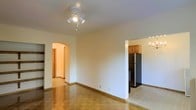 This single-floor residence offers a welcoming and functional layout with classic design details throughout. The main living area is characterized by warm parquet wood flooring that adds a timeless charm to the space, while white walls and a ceiling-mounted multi-bulb light fixture provide a bright and airy atmosphere.

A graceful arched doorway leads from the living area into the hallway, adding an elegant architectural touch that enhances the overall character of the home. This arched passage connects the living space to the rest of the residence in a seamless and visually appealing way.

Adjacent to the living area, the kitchen is accessible through an open entryway and features wood cabinetry in a warm honey tone, offering ample storage. A stainless steel refrigerator anchors the kitchen, lending a modern and functional element to the space. A wall-mounted light fixture in the kitchen area adds a decorative accent while providing additional illumination.

Throughout the space, clean white baseboards and neutral wall tones create a cohesive and understated aesthetic that allows for versatile furnishing and decorating. The overall ambiance of the residence is one of comfort and practicality, with architectural details that give the home a distinctive and inviting character.