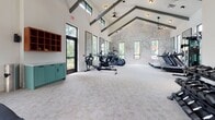 Located at 8400 Palmetto Commerce Pkwy in Ladson, SC, this single-floor fitness center is designed to deliver a premium workout experience. The space is defined by soaring vaulted ceilings accented with dark exposed beams, creating an airy, open atmosphere throughout. Large black-framed windows line the walls, flooding the interior with natural light and offering views of the surrounding greenery outside.

The cardio zone features a row of treadmills, elliptical trainers, and a rowing machine arranged along one side of the facility, while ceiling fans keep the space comfortable during intense sessions. A striking topographic map mural serves as a bold focal point on one wall, adding a distinctive design element to the environment.

The strength training area is anchored by a vivid orange power rack, accompanied by medicine balls, BOSU trainers, barbells, and a full dumbbell rack. Large wall-mounted mirrors enhance the sense of space and allow users to monitor their form. A dedicated free weights and functional training zone provides ample room for floor-based exercises and stretching.

Built-in cabinetry in teal and warm wood tones offers practical storage and adds a refined, intentional aesthetic to the space. The light neutral carpet throughout ties the design together, balancing the energetic equipment with a clean, welcoming environment suited for a wide range of fitness activities.