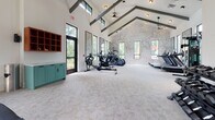 Located at 8400 Palmetto Commerce Pkwy in Ladson, SC, this single-floor fitness center is designed to deliver a premium workout experience. The space is defined by soaring vaulted ceilings accented with dark exposed beams, creating an airy, open atmosphere throughout. Large black-framed windows line the walls, flooding the interior with natural light and offering views of the surrounding greenery outside.

The cardio zone features a row of treadmills, elliptical trainers, and a rowing machine arranged along one side of the facility, while ceiling fans keep the space comfortable during intense sessions. A striking topographic map mural serves as a bold focal point on one wall, adding a distinctive design element to the environment.

The strength training area is anchored by a vivid orange power rack, accompanied by medicine balls, BOSU trainers, barbells, and a full dumbbell rack. Large wall-mounted mirrors enhance the sense of space and allow users to monitor their form. A dedicated free weights and functional training zone provides ample room for floor-based exercises and stretching.

Built-in cabinetry in teal and warm wood tones offers practical storage and adds a refined, intentional aesthetic to the space. The light neutral carpet throughout ties the design together, balancing the energetic equipment with a clean, welcoming environment suited for a wide range of fitness activities.