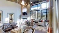 This sophisticated single-floor space makes an immediate impression with its dramatic double-height ceilings that create an expansive, airy atmosphere. A statement fireplace wall clad in sleek metallic paneling anchors the main lounge area, with a wall-mounted television above and a modern linear fireplace below, serving as a striking architectural focal point. Flanking this feature wall, floor-to-ceiling windows flood the interior with abundant natural light while offering open views of the surrounding streetscape, seamlessly connecting the indoors with the outside environment. The ceiling itself adds visual interest with its rich, dark metallic finish, contrasted by a cascading chandelier that provides warm, ambient illumination. The lounge is thoughtfully appointed with a curated mix of contemporary furnishings, including a large sectional sofa, tufted accent chairs in brushed metallic finishes, and leather seating arrangements that invite relaxed conversation. Layered area rugs in muted, multi-tonal patterns define the seating zones atop hardwood-style flooring, adding warmth and texture. The overall ambiance is one of upscale, modern comfort — a well-designed communal space that balances bold architectural elements with inviting, livable details.