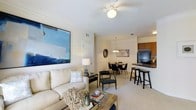 This single-floor residence is designed with an open-concept layout that connects the living, dining, and kitchen areas into one cohesive and functional space. The living area features plush carpeted flooring, a ceiling fan with a central light fixture, and ample room for comfortable seating. A large abstract canvas painting in cool blue tones serves as a striking focal point on the wall, complementing the neutral color palette throughout.

The dining area sits adjacent to the living space and is illuminated by an elegant chandelier, creating an inviting atmosphere for meals and gatherings. A round dining table surrounded by sleek black chairs anchors this area, which transitions naturally into the kitchen.

The kitchen is defined by warm wood cabinetry, dark countertops, and a breakfast bar with bar-height seating, offering both functionality and a casual dining option. Black appliances, including a full-sized refrigerator, provide a bold contrast to the lighter tones of the surrounding space.

Throughout the residence, the consistent use of neutral walls, soft carpeting, and carefully chosen lighting creates a warm and welcoming ambiance. The layout is efficient and thoughtfully designed, making excellent use of the available space while maintaining an airy, open feel.