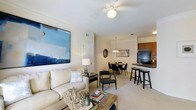 This single-floor residence is designed with an open-concept layout that connects the living, dining, and kitchen areas into one cohesive and functional space. The living area features plush carpeted flooring, a ceiling fan with a central light fixture, and ample room for comfortable seating. A large abstract canvas painting in cool blue tones serves as a striking focal point on the wall, complementing the neutral color palette throughout.

The dining area sits adjacent to the living space and is illuminated by an elegant chandelier, creating an inviting atmosphere for meals and gatherings. A round dining table surrounded by sleek black chairs anchors this area, which transitions naturally into the kitchen.

The kitchen is defined by warm wood cabinetry, dark countertops, and a breakfast bar with bar-height seating, offering both functionality and a casual dining option. Black appliances, including a full-sized refrigerator, provide a bold contrast to the lighter tones of the surrounding space.

Throughout the residence, the consistent use of neutral walls, soft carpeting, and carefully chosen lighting creates a warm and welcoming ambiance. The layout is efficient and thoughtfully designed, making excellent use of the available space while maintaining an airy, open feel.