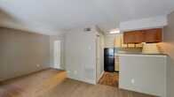 This single-floor studio offers a practical, open-concept layout that efficiently combines living and functional spaces. The main living area is finished with wall-to-wall carpeting in a neutral tan tone, creating a cohesive and understated aesthetic throughout. Large windows along the front wall allow natural light to flow freely into the space, offering views of the surrounding outdoor greenery and providing a bright, airy atmosphere. A front entry door is flanked by these windows, making the entryway feel welcoming and well-lit.

The kitchen is semi-enclosed, set slightly apart from the living area by a partial wall that serves as a natural divider. It features wood-toned cabinetry in both light and medium finishes, providing ample upper storage. The kitchen is equipped with a black refrigerator, a stainless steel single-basin sink with laminate countertops, and under-cabinet lighting. Wood-look flooring runs through the kitchen, distinguishing it from the carpeted living zone.

A closet with louvered doors provides convenient storage within the main living space. The bathroom includes a full-size soaking tub with a tiled surround in classic white, complemented by chrome fixtures. The textured ceiling is consistent throughout, adding a traditional residential character to the overall space.
