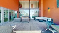 This contemporary outdoor terrace is set against a striking building facade clad in bold terracotta-orange paneling, creating a vibrant architectural backdrop. The space is divided into several distinct zones, each designed for a different social function. A central lounge area anchors the terrace, featuring woven rattan seating arranged around a linear fire pit table, providing a warm and inviting atmosphere for gatherings. Adjacent to this, a curved sectional sofa paired with a round concrete coffee table offers an additional relaxed seating option.

Along the terrace wall, a mounted flat-screen television with integrated wall sconces creates a dedicated outdoor viewing area, complemented by a modern sofa and low concrete table. Decorative planters add subtle greenery to the space. A separate dining zone features multiple round tables with woven chairs, positioned near an open railing that frames unobstructed views of the surrounding tree line and streetscape, allowing ample natural light throughout the day.

Floor-to-ceiling glass walls and sliding doors connect the terrace to the interior, reinforcing a seamless indoor-outdoor flow. The polished concrete flooring throughout ties the zones together cohesively, making this terrace a well-appointed, multifunctional outdoor space.