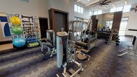 This well-appointed fitness center is designed to accommodate a full range of workout needs within a single, open floor plan. The space is characterized by soaring ceilings with clerestory windows positioned along the upper walls, flooding the interior with natural light while maintaining privacy. Large floor-to-ceiling windows along the perimeter further enhance the bright, airy atmosphere and offer views of the surrounding outdoors.

The flooring transitions between durable wood-look planks in the dedicated stretching and studio area and patterned, multi-tone carpet tiles throughout the main workout floor. Full-length wall mirrors are strategically placed to visually expand the space and assist with exercise form. Ceiling fans and contemporary lighting fixtures, including recessed and pendant-style cluster lights, ensure consistent illumination and airflow.

The strength training section is equipped with a comprehensive array of Hoist-brand cable and selectorized machines, a multi-station functional trainer, and a well-stocked dumbbell rack. A dedicated cardio zone features Cybex treadmills, ellipticals, and upright bikes. Stability balls, resistance bands, and yoga mats are neatly organized near a fitness-themed accent wall. Glass-panel doors connect the fitness center to an adjacent lounge or common area, reinforcing the facility's open, community-oriented design.