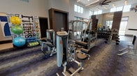 This well-appointed fitness center is designed to accommodate a full range of workout needs within a single, open floor plan. The space is characterized by soaring ceilings with clerestory windows positioned along the upper walls, flooding the interior with natural light while maintaining privacy. Large floor-to-ceiling windows along the perimeter further enhance the bright, airy atmosphere and offer views of the surrounding outdoors.

The flooring transitions between durable wood-look planks in the dedicated stretching and studio area and patterned, multi-tone carpet tiles throughout the main workout floor. Full-length wall mirrors are strategically placed to visually expand the space and assist with exercise form. Ceiling fans and contemporary lighting fixtures, including recessed and pendant-style cluster lights, ensure consistent illumination and airflow.

The strength training section is equipped with a comprehensive array of Hoist-brand cable and selectorized machines, a multi-station functional trainer, and a well-stocked dumbbell rack. A dedicated cardio zone features Cybex treadmills, ellipticals, and upright bikes. Stability balls, resistance bands, and yoga mats are neatly organized near a fitness-themed accent wall. Glass-panel doors connect the fitness center to an adjacent lounge or common area, reinforcing the facility's open, community-oriented design.