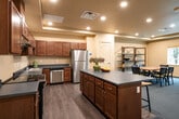 This single-floor community facility is thoughtfully designed to accommodate a variety of group activities and shared living needs. The fully equipped kitchen serves as a central hub, featuring rich dark cherry wood cabinetry, dark blue countertops, stainless steel appliances including a refrigerator, dishwasher, and range, and durable hardwood-style flooring. A central island with additional counter space enhances functionality and accessibility. Adjacent to the kitchen, an open dining area provides ample seating around a solid wood table, complemented by open shelving units and a window offering views of surrounding greenery. The lounge area is carpeted in deep blue and furnished with multiple round tables and mixed seating arrangements, creating a relaxed, social atmosphere. A separate TV room features plush dark leather armchairs with decorative pillows, a wall-mounted flat-screen television, and windows that bring in natural light. Framed artwork adorns the walls throughout. Recessed lighting and tray ceilings are consistent throughout the space, contributing to a warm, inviting ambiance. The layout is open and accessible, with clearly defined zones that support both communal interaction and quieter, individual use.