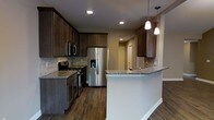 This single-story space showcases a well-appointed kitchen as its centerpiece. The kitchen features rich, dark espresso-toned shaker-style cabinetry that extends to the ceiling, providing generous storage throughout. The cabinetry is complemented by granite countertops with subtle grey and beige toning, adding a refined, natural element to the design.

A peninsula with a white base extends into the adjoining area, topped with matching granite and illuminated by two sleek pendant lights, making it ideal for casual dining or meal prep. The stainless steel appliance suite includes a gas range, built-in microwave, and a French door refrigerator with a water and ice dispenser, offering a cohesive and functional cooking environment.

A stone tile backsplash runs along the cooking wall, adding texture and visual interest between the counters and upper cabinets. Recessed lighting provides consistent, even illumination throughout the kitchen. Dark hardwood flooring grounds the space and ties together the warm tones of the cabinetry. A hallway visible in the background suggests additional rooms beyond the kitchen area.