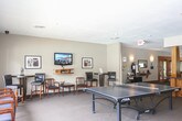 This single-floor community amenity space is thoughtfully designed to provide residents with a variety of recreational and social areas. The lounge area features warm neutral-toned walls, carpeted flooring, and comfortable seating arrangements including wood-framed chairs with cushioned seats and high-top bistro tables with bar-height chairs. A wall-mounted flat-screen television sits above floating wooden shelving, making the space ideal for relaxed viewing. Framed artwork decorates the walls throughout, adding a polished, welcoming atmosphere.

The game and activity room offers generous open space, highlighted by a full-size ping-pong table positioned beneath recessed lighting. Large sliding glass doors flood the room with natural light and provide direct views of an outdoor pool area, creating a seamless indoor-outdoor connection. A cozy accent chair and decorative plants contribute to the inviting ambiance.

Adjacent to the game area, a fitness room is visible through glass partitions, equipped with exercise machines and illuminated by overhead lighting. The common area also includes a reception or service counter with warm wood cabinetry and dark countertops, along with a self-serve coffee station nearby. Corridors connect the various zones, ensuring easy navigation throughout this versatile and well-equipped community facility.