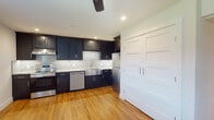 This single-floor residence offers a clean, modern aesthetic complemented by warm hardwood flooring that flows consistently throughout the entire space. The kitchen serves as a standout centerpiece, showcasing rich dark shaker-style cabinetry paired with light granite countertops and a striking mosaic tile backsplash. A deep stainless steel farmhouse sink, stainless steel appliances — including a refrigerator, dishwasher, and range — and a full-height pantry with paneled white doors add both functionality and style. Recessed lighting and a ceiling fan keep the kitchen bright and comfortable. The open layout extends into a generously sized living area, which is bathed in natural light through a window overlooking greenery, and is further enhanced by a ceiling fan with integrated lighting. An additional room, ideal as a bedroom or flex space, mirrors the same warm flooring and neutral tones, featuring a window with blinds and a large paneled closet. The bathroom is elegantly finished with white subway tile surrounding a soaking tub and shower combination, accented by a hexagonal mosaic tile niche, dark grout detailing, and granite countertops — creating a cohesive, refined look throughout the home.