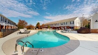 Located at 10035 Dabney Dr in Charlotte, North Carolina, this outdoor amenity area serves a multi-building residential community and is designed to provide a welcoming and functional recreational environment. The centerpiece is a generously sized freeform swimming pool with clear turquoise water, gently curved edges, and designated depth markers. The pool is flanked by a broad concrete sundeck furnished with multiple lounge chairs and side tables, offering ample space for sunbathing and relaxation. A wrought iron perimeter fence encloses the pool area, providing both security and a defined boundary.

Adjacent to the pool, a single-story clubhouse building features large floor-to-ceiling glass panels and double glass entry doors framed in aluminum, allowing natural light to flow freely into the interior. The exterior of the clubhouse is finished with a combination of light siding and classic brick detailing, consistent with the architectural character of the surrounding residential buildings. These multi-story brick buildings with dark shuttered windows form a cohesive backdrop around the pool courtyard, enhancing the sense of an enclosed, community-focused setting. Mature trees and seasonal landscaping complete the outdoor environment, contributing to a comfortable and inviting atmosphere under open North Carolina skies.