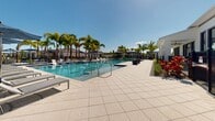 Located at 4180 Yardly Dr in Palmetto, Florida, this single-level outdoor amenity space delivers a resort-style experience defined by thoughtful design and lush Florida landscaping. At the heart of the space is a generously sized swimming pool with clear blue water, framed by wide, light-toned paver decking and flanked by rows of lounge chairs — ideal for sunbathing beneath the Florida sun. Tall, mature palm trees are positioned throughout the grounds, providing natural shade and reinforcing the tropical ambiance.

A series of bold navy blue canopy structures line one side of the pool area, offering covered seating and refuge from direct sunlight, while freestanding umbrellas provide additional shade around the perimeter. Ornamental grasses, colorful shrubs, and layered plantings with decorative gravel accents create well-maintained garden beds that add texture and visual interest throughout.

A dedicated lawn area features a hammock suspended between palm trees, offering a quiet retreat for relaxation. Outdoor dining areas with umbrella-shaded tables and chairs provide communal gathering spots, enhanced by an ornamental iron fence and gated entry. The clubhouse building features a clean, contemporary exterior with large windows and coordinated landscaping. The entire space is bathed in natural light and open to the sky, creating a vibrant, welcoming atmosphere suited for community recreation and leisure.