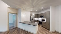 Located at 6004 Grandover Village Rd in Greensboro, NC, this single-floor residence offers a thoughtfully designed open-concept layout with contemporary finishes throughout. The kitchen serves as a central highlight, featuring taupe shaker-style cabinetry extending to the ceiling, stainless steel appliances including a French door refrigerator with ice maker, electric range, and built-in microwave. A peninsula with a dark granite countertop and undermount sink provides additional workspace and seating, illuminated by elegant glass pendant lights and recessed ceiling fixtures. Wood-look plank flooring flows throughout the main living areas, adding warmth and cohesion to the open floor plan. The living area is generously proportioned with clean white walls and easy access to the kitchen. Bedrooms are carpeted for comfort; the primary bedroom benefits from natural light through a large window, a ceiling fan, and direct access to a walk-in closet. The primary bathroom offers a granite-topped vanity with dark cabinetry, a tub-shower combination, and tile flooring. A second bathroom mirrors the refined aesthetic with similar finishes and ample storage. The overall design is clean, modern, and move-in ready, reflecting a well-maintained and cohesively styled living environment.