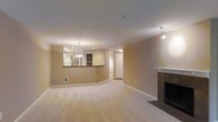 This single-floor residence offers a well-designed, open-concept layout that maximizes the sense of space and flow throughout the interior. The expansive living area is finished with plush wall-to-wall carpeting in a neutral tone, creating a warm and comfortable atmosphere. A classic fireplace with a clean, white mantle serves as a natural focal point, anchoring the living space with both style and function. Recessed lighting and a decorative pendant light fixture illuminate the room evenly, complementing the warm, neutral wall tones that carry throughout the space. A half-wall pass-through connects the living area to the kitchen, which features built-in open shelving and a refrigerator visible from the main living space. The pendant light above the kitchen counter adds a touch of elegance to the cooking area. A doorway leads to additional rooms beyond, suggesting a practical and well-organized floor plan. The overall ambiance is calm, bright, and inviting, with a cohesive color palette of soft beige and white tones that lend the space a timeless, move-in-ready appeal suitable for a variety of living arrangements.