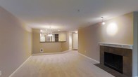 This single-floor residence offers a well-designed, open-concept layout that maximizes the sense of space and flow throughout the interior. The expansive living area is finished with plush wall-to-wall carpeting in a neutral tone, creating a warm and comfortable atmosphere. A classic fireplace with a clean, white mantle serves as a natural focal point, anchoring the living space with both style and function. Recessed lighting and a decorative pendant light fixture illuminate the room evenly, complementing the warm, neutral wall tones that carry throughout the space. A half-wall pass-through connects the living area to the kitchen, which features built-in open shelving and a refrigerator visible from the main living space. The pendant light above the kitchen counter adds a touch of elegance to the cooking area. A doorway leads to additional rooms beyond, suggesting a practical and well-organized floor plan. The overall ambiance is calm, bright, and inviting, with a cohesive color palette of soft beige and white tones that lend the space a timeless, move-in-ready appeal suitable for a variety of living arrangements.