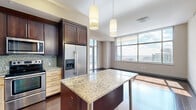 This three-bedroom, two-bathroom high-rise residence combines contemporary design with functional living across a well-proportioned single floor. The open-concept kitchen and dining area serves as the heart of the home, featuring rich dark wood cabinetry, light maple lower cabinets, granite countertops, a mosaic tile backsplash, and a full suite of stainless steel appliances including a French door refrigerator, electric range, microwave, and dishwasher. A granite-topped island with pendant lighting anchors the space, which flows seamlessly into the dining area framed by floor-to-ceiling windows with sweeping city views. The hallway is distinguished by track lighting and a built-in niche with a marble ledge, adding an architectural accent. The bedrooms are carpeted and bathed in natural light through large city-view windows, with five-panel gray doors and ample closet space supported by wire shelving systems. The bathrooms feature granite vanities with undermount sinks, dark wood cabinetry, and large-format tile flooring. A dedicated laundry room houses a full-size washer and dryer with additional wire shelving. Rich hardwood floors run throughout the main living areas, completing this polished urban residence.