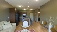 This single-floor residence showcases a thoughtfully designed open-concept layout that integrates the kitchen, dining, and living areas into one cohesive and functional space. The kitchen is equipped with dark wood cabinetry, granite countertops, stainless steel appliances, and a black refrigerator, all anchored by a central island with bar seating — ideal for casual dining or entertaining. Recessed and flush-mount lighting throughout ensures the space is well-lit and inviting. The living area flows naturally from the kitchen, featuring light hardwood-style flooring that extends throughout the main space, adding warmth and continuity. A vibrant patterned area rug and contemporary seating, including a white accent chair, bring a modern touch to the room. Warm olive-toned walls complement the rich cabinetry, creating a cohesive and grounded color palette. A hallway visible from the main area leads to additional rooms, including what appears to be a bathroom, suggesting a well-structured floor plan beyond the open living space. The overall ambiance is modern, comfortable, and practical, offering a welcoming environment suited for everyday living.