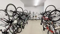 This single-level bike storage room is a practical, well-organized space designed to house a substantial number of bicycles. The room features clean white walls and a light-toned floor, creating a bright and functional environment illuminated by overhead fluorescent strip lighting. Wall-mounted bicycle hooks line the back wall, allowing bikes to be hung vertically to maximize space efficiency. In addition to the wall-mounted racks, additional bicycles are stored along both sides of the room using floor-level angled rack systems, enabling a high-density storage arrangement. The layout creates a clear central corridor, providing easy access to all stored bicycles. The variety of bikes present — including road bikes, commuter bikes, and others — reflects the room's capacity to serve a diverse range of users. The interior finish is minimal and utilitarian, with smooth walls and durable flooring suited to the demands of a shared storage facility. Overall, the space is thoughtfully designed to prioritize practicality and capacity, making it a highly functional amenity for cyclists.