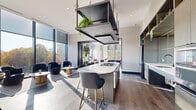 This thoughtfully designed single-floor space seamlessly blends a fully equipped kitchen with generous communal lounge areas, creating an environment suited for both casual gatherings and collaborative use. The open-concept kitchen serves as a central focal point, anchored by a curved white quartz island with an integrated gas cooktop and stylish bar seating. A striking suspended black metal rack overhead adds architectural drama while incorporating live greenery. The kitchen is further equipped with stainless steel appliances, a deep sink, built-in wall ovens, and ample cabinetry with open shelving displaying curated décor.

The lounge areas extend generously from the kitchen, defined by wide-plank hardwood flooring throughout. Floor-to-ceiling windows wrap around the perimeter, inviting abundant natural light and offering expansive outdoor views. A large sectional sofa anchors one seating area beneath a sculptural pendant chandelier, while a separate lounge nook features warm tan leather sofas, bold artwork, and built-in bookshelves styled with books and decorative objects. Vibrant geometric area rugs, lush indoor plants, and a carefully curated mix of modern furnishings contribute to an inviting, upscale ambiance throughout the space.