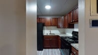 This single-floor unit offers a practical and well-appointed layout with consistent updates throughout. The kitchen is a functional standout, featuring rich dark cherry cabinetry paired with light granite-style countertops and a white subway tile backsplash. Black appliances, including a coil-burner range and refrigerator, complement the cabinetry, while light vinyl plank flooring ties the space together cleanly.

The living area is open and versatile, finished with the same light wood-tone vinyl plank flooring and neutral taupe walls. A wall-mounted HVAC unit provides climate control, and a window allows natural light to filter into the room. The bedroom offers a similarly neutral palette with a built-in closet featuring bifold doors and ample shelving, providing practical storage. The flooring transitions to a slightly warmer wood tone in this space.

The bathroom is efficiently laid out, featuring a white vanity with under-cabinet storage, a wall-mounted medicine cabinet with lighted mirror, and a tub-shower combination accented by distinctive teal ceramic tile surround. Chrome fixtures and grab bars add functionality. Throughout the unit, panel-style ceilings and crisp white baseboards create a clean, cohesive aesthetic.