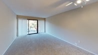 This single-floor, 2-bedroom, 1.5-bathroom residence features a well-organized layout designed for comfortable everyday living. The open living area is carpeted and bathed in natural light from a large sliding glass door that leads to a private balcony with outdoor views, complemented by vertical blinds for adjustable light control. A ceiling fan adds to the comfort of the space. The kitchen is positioned adjacent to the living area in an open-concept fashion, featuring warm wood-toned cabinetry both above and below the counter, a white electric coil range with an overhead exhaust hood, and a white refrigerator, providing efficient use of the available space. Both bedrooms are carpeted and offer ample closet storage — one bedroom includes a sliding mirrored closet with a hanging rod, while the other features a standard closet alongside a built-in shelving unit visible in the hallway. The full bathroom includes a white vanity with cabinet storage below, a medicine cabinet with mirror above, and tile flooring in a warm neutral tone. The textured ceilings and neutral warm-beige wall tones throughout create a cohesive and understated aesthetic across the entire residence.