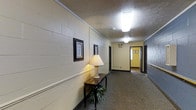 This single-story space presents a long interior hallway that serves as a central circulation corridor. The walls are constructed of painted concrete block, finished in a creamy off-white tone that reflects light and contributes to a clean, institutional aesthetic. A chair rail molding runs along the lower portion of the walls, adding a subtle architectural detail that breaks up the vertical plane.

The flooring throughout the corridor is covered in a neutral, low-pile carpet in a muted gray-brown tone, providing durability and sound absorption along the length of the passageway. Overhead, a textured ceiling houses flush-mounted fluorescent light fixtures that provide consistent, even illumination down the hall.

Decorative touches soften the utilitarian character of the space. A narrow wooden console table is positioned along one wall, topped with a classic table lamp that adds warm ambient light. Framed botanical artwork adorns the walls, contributing a welcoming, finished feel. At the far end of the corridor, a yellow-painted wall and additional framed pieces are visible, along with multiple doorways branching off in both directions, suggesting several rooms or suites beyond.