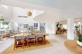 Located at 1155 SE Seventh Ave in Dania Beach, Florida, this two-story space offers a generous and thoughtfully designed layout spread across a spacious main floor and a smaller upper level. The architecture is defined by dramatic double-height ceilings, crisp white walls, and large-format light tile flooring that unifies the expansive ground floor.

The main level is the heart of the space, featuring an open-plan area that accommodates multiple functional zones, including communal gathering areas with large wooden tables and chairs, a kitchen equipped with stainless steel appliances and light wood cabinetry, and a dedicated workspace with individual desks and ample natural light from multiple windows. Glass-panel partitions with black metal framing define a private conference area without compromising the open feel. Woven pendant lighting and tropical plants add warmth to the otherwise clean, minimalist aesthetic. Views of an outdoor pool area are visible through large windows and glass doors.

The upper level features a bright, open loft-style area with vaulted ceilings, a striking circular window overlooking lush greenery, and a black metal railing overlooking the floor below. This level provides a quieter, more intimate setting with seating and lounge furnishings.