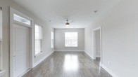This well-appointed single-floor space offers a clean, modern aesthetic defined by neutral tones and thoughtful design details. The main living area is finished with smooth, light gray hardwood-style flooring that reflects natural light beautifully, creating a warm and inviting atmosphere. Crisp white walls and bright white baseboards and door trim give the space a polished, cohesive look throughout.

Multiple windows fitted with classic white horizontal blinds allow generous amounts of natural light to fill the room, enhancing the open and airy feel. A ceiling fan with a built-in light fixture is centrally positioned overhead, providing both comfort and functional illumination. The ceiling is smooth and clean, maintaining the minimalist character of the space.

An open doorway leads to an adjacent room or hallway, suggesting a practical and connected floor plan. The overall ambiance is fresh, bright, and move-in ready, with a neutral palette that offers flexibility for a variety of interior styles. The combination of durable flooring, ample natural light, and clean architectural lines makes this a highly functional and visually appealing space.