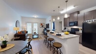 This thoughtfully designed single-floor residence offers a seamless open-concept layout that connects the kitchen, dining, and living areas into one cohesive, functional space. The kitchen is a standout feature, equipped with dark espresso cabinetry, granite-style countertops, black appliances including a gas range, microwave, and refrigerator, and a peninsula island with bar seating illuminated by pendant lighting. A dedicated in-unit laundry closet with a full-size washer and dryer is conveniently accessible from the kitchen corridor. The dining area accommodates a round table with modern seating and is accented by a bold piece of wall art, creating an intimate yet stylish setting. The living room is warm and inviting, with light wood-style flooring, a set of French doors that bring in natural light, and ample space for comfortable furnishings. The bedroom features plush carpeting, large windows with blinds and drape panels for privacy, and generous storage. The full bathroom is equipped with grab bars for accessibility, a vanity with dark cabinetry, and a tub-shower combination. Throughout the residence, clean white walls and consistent finishes contribute to a bright, modern, and cohesive aesthetic.