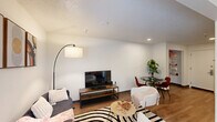 Located at 123 E McCullough Dr in Charlotte, NC, this single-floor unit offers a thoughtfully organized layout that maximizes its compact footprint. The open living area serves as the heart of the space, featuring warm hardwood-style flooring, neutral walls, and a textured ceiling that adds subtle character. Natural light filters in through a window fitted with blinds, brightening the room and creating an inviting atmosphere. A wall-mounted HVAC unit ensures year-round comfort. The living area is versatile enough to accommodate both a lounging and dining arrangement, with a defined alcove section providing a cozy nook ideal for a daybed or additional seating. Recessed lighting and an open pass-through to a kitchenette area enhance the sense of connectivity throughout the space. The full bathroom is cleanly finished with white tile surrounding a shower-tub combo, complete with a safety grab bar. Wood-look flooring continues into the bathroom, tying the aesthetic together. A vanity with a white countertop sink, a toilet, and a freestanding metal shelving unit provide practical storage and functionality. Overall, the unit presents a clean, modern aesthetic well-suited for efficient, comfortable living in the University City area of Charlotte.