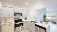 This well-appointed single-floor residence is designed with a clean, contemporary aesthetic throughout. The heart of the home is a thoughtfully designed kitchen, featuring crisp white shaker-style cabinetry with brushed nickel hardware, bright white countertops, and a full suite of stainless steel appliances including a side-by-side refrigerator, electric range, built-in microwave, and dishwasher. A peninsula with a double-basin sink provides additional prep space and separates the kitchen from the adjoining living area, which is illuminated by a modern multi-arm chandelier. Light wood-style flooring extends through the main living areas, contributing to the space's airy, open feel. Recessed lighting throughout ensures consistent, well-distributed illumination. A glass-panel door with window blinds opens to an outdoor area, allowing natural light to flow into the living space. The bedroom offers plush carpeting, a generous double-door closet, and a tray ceiling detail that adds architectural character. The overall design palette — neutral walls, white trim, and light finishes — creates a cohesive, bright, and inviting atmosphere throughout the entire residence.