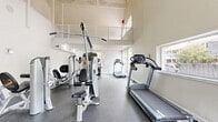 This two-story fitness center is thoughtfully designed to accommodate a full range of workout activities across two distinct levels. The main floor serves as the primary exercise area, featuring durable rubber flooring and a generous layout that comfortably houses an array of cardio and strength-training equipment, including treadmills, stationary bikes, cable machines, and multi-use weight stations. Recessed lighting and large windows along the walls allow natural light to fill the space, creating a bright and energizing atmosphere. Soaring ceilings with exposed beam detailing lend the main floor a sense of openness, while a bold yellow ceiling accent adds a vibrant pop of color near the staircase entry. A staircase leads to the upper mezzanine level, which is enclosed by a white metal mesh railing and offers an open, airy feel thanks to vaulted, pitched ceilings and wide windows overlooking the surrounding neighborhood. This upper level features light hardwood flooring and serves as a dedicated flex or yoga studio, complete with exercise mats and balance equipment. The overall design balances functionality with a clean, modern aesthetic throughout both floors.