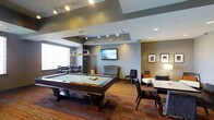 This single-floor amenity lounge is thoughtfully designed to offer a well-rounded recreational and social experience. At the heart of the space sits a full-size pool table, illuminated by a sleek pendant light fixture that serves as a visual focal point. The room features a multi-toned, striped carpet that adds warmth and texture to the flooring, complementing the contemporary color palette of gray, taupe, and warm earth tones that define the walls and ceiling design. A wall-mounted flat-screen TV is centrally positioned, making it accessible from multiple seating areas throughout the room. The lounge is divided into distinct zones: a gaming area centered around the pool table, a casual seating arrangement with upholstered chairs and a sofa accented with throw pillows, and a dining or meeting area with a table and chairs near a frosted glass window that allows diffused natural light to filter in. Recessed and track lighting throughout the space ensures even, well-distributed illumination. Framed artwork on the walls adds a refined touch, while the overall layout encourages both lively group interaction and quiet relaxation. The space strikes a balance between modern sophistication and inviting comfort.