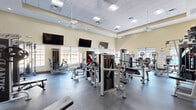 Located at 9000 Colby Dr in Fort Myers, Florida, this dedicated fitness center offers a generously sized, single-floor exercise room designed to accommodate a full range of workout routines. The space features soaring ceilings that lend an open, airy atmosphere, complemented by warm neutral-toned walls and durable rubber flooring suited for high-traffic athletic use. Natural light filters in through multiple large windows fitted with roller shades, offering views of the outdoor pool area and surrounding Florida landscaping. Multiple wall-mounted flat-screen televisions are positioned throughout the room for entertainment during workouts. The equipment selection is extensive, including a row of treadmills and an elliptical machine along the windowed wall, as well as a wide variety of cable and selectorized strength machines, free weight benches, and a dedicated free weight rack with mirrors. Ceiling fans and a recessed lighting grid with a drop-tile ceiling ensure comfort and visibility across the entire floor. Warm wood-trimmed double doors provide access points on multiple sides of the room. The overall ambiance is clean, modern, and functional, making this fitness center a well-equipped facility for residents at this Fort Myers location.