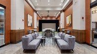 This upscale amenity space presents a thoughtfully designed, multi-functional environment that seamlessly blends social lounging, event hosting, and recreational entertainment. The space is anchored by soaring ceilings and warm terracotta-toned textured wall panels that lend a rich, contemporary character throughout.

The lounge area features plush, grey upholstered sofas and armchairs arranged around sleek round side tables, creating intimate conversation groupings. Herringbone-patterned tile flooring transitions into geometric carpet zones, adding visual depth and defining different functional areas. Large-scale contemporary artwork punctuates the walls, contributing color and artistic energy to the interior.

A central high-top communal table with bar-height seating serves as a collaborative workspace or social gathering hub. Floor-to-ceiling glass walls on one side open the space visually to an adjacent lobby or common area, enhancing the sense of openness and connectivity.

At the rear of the space, a dedicated golf simulator bay features synthetic green turf flooring, black curtain surround, and a large projection screen — offering an immersive recreational experience. A ceiling-mounted projector serves the broader room, making the space equally suited for presentations or screenings. Recessed lighting and wall sconces provide warm, layered illumination throughout, completing the polished and inviting atmosphere.