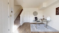 This three-floor home is thoughtfully designed to offer a variety of functional living spaces across its levels. The entry level features wood-look hardwood-style flooring that carries throughout much of the main living areas, establishing a warm and cohesive aesthetic. A dedicated home office area on this level includes ample space for a glass-top desk and seating, with neutral walls and natural light creating a productive environment.

The kitchen is outfitted with warm honey-toned cabinetry, stainless steel appliances, a black refrigerator, and grey countertops, complemented by a peninsula that adds extra prep and serving space. Adjacent to the kitchen, the dining area benefits from large windows dressed in sheer curtains that invite generous natural light, centering around a round wood dining table.

The carpeted living room features elevated ceilings with a tray detail, lending a sense of openness. The bedroom is carpeted and well-lit by a large window with blinds, offering a peaceful retreat with closet storage. The bathroom includes dual sinks, warm wood cabinetry, grey countertops, and wood-look flooring. Carpeted stairs with wood-stained railings connect all three floors seamlessly.