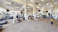 Located in Johns Creek, GA, this generously sized fitness center spans a single floor and delivers a bright, welcoming environment for a comprehensive workout experience. The space is defined by soaring coffered ceilings accented with recessed lighting and large ceiling fans, creating excellent airflow and an open, airy atmosphere throughout.

The exercise floor is thoughtfully organized into distinct training zones. A dedicated cardio area lines the perimeter beneath floor-to-ceiling windows, offering a row of treadmills and stationary bikes with direct views of lush outdoor greenery and a pool area — an energizing backdrop for any workout. Strength training is well-represented with a wide array of Life Fitness selectorized machines, free weight stations, and cable systems arranged across patterned, durable carpet flooring interspersed with rubber mat zones for safety and versatility.

Full-length mirrors span key wall sections, visually expanding the space and allowing users to monitor their form. Wall-mounted flat-screen televisions are positioned throughout for entertainment during exercise. The neutral color palette of warm whites and beige tones, combined with abundant natural light pouring in through multiple large window banks, creates a clean and motivating atmosphere. The overall design balances functionality with an upscale aesthetic, making this facility a standout amenity.